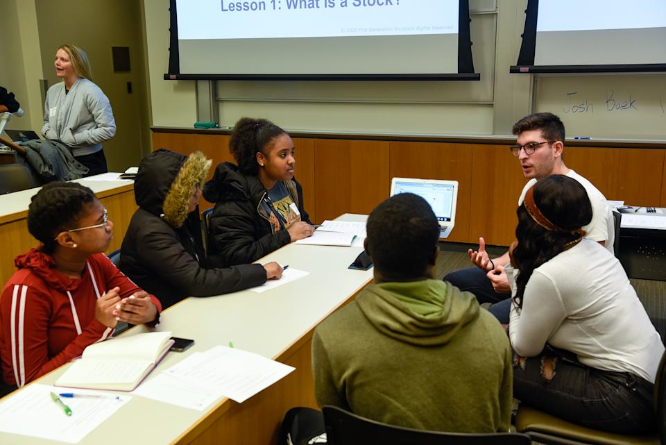 High school students gathered around a table discuss stock investing.